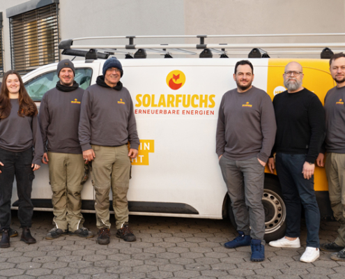 Auf dem Foto ist das Team von Solarfuchs, acht Personen, vor einem weißen Fahrzeug mit dem Logo "SOLARFUCHS" zu sehen. Das Fahrzeug trägt die Aufschrift "Erneuerbare Energien". Alle Personen sind in grauen Sweatshirts und beige Hosen gekleidet. Einige tragen auch Mützen oder Hüte. Sie stehen in einer Reihe, teilweise leicht nach links oder rechts gedreht.
