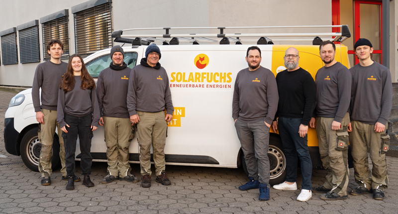 Auf dem Foto ist das Team von Solarfuchs, acht Personen, vor einem weißen Fahrzeug mit dem Logo "SOLARFUCHS" zu sehen. Das Fahrzeug trägt die Aufschrift "Erneuerbare Energien". Alle Personen sind in grauen Sweatshirts und beige Hosen gekleidet. Einige tragen auch Mützen oder Hüte. Sie stehen in einer Reihe, teilweise leicht nach links oder rechts gedreht.
